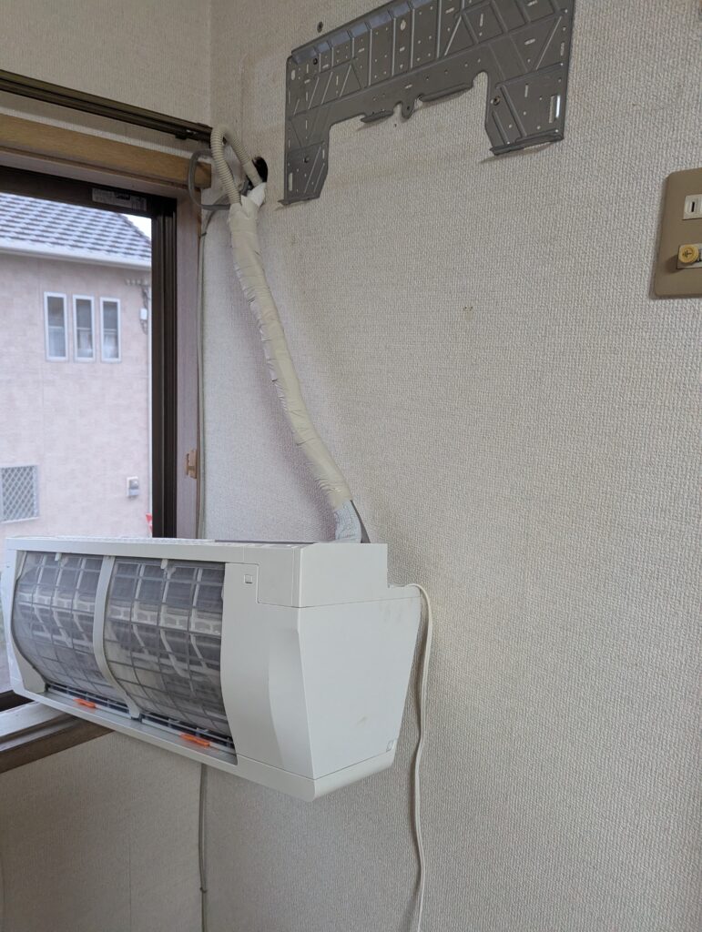 配管の束を左上の壁穴へ通し、室内機を窓枠に仮置きして持ち上げようとしている様子